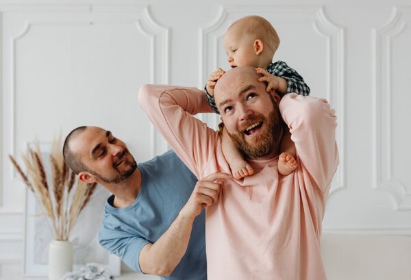 Ein Vater trägt Baby auf den Schultern, zweiter Vater steht daneben und lächelt es an