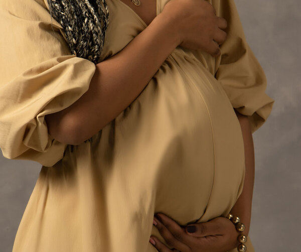 Schwangere Frau im ockerfarbenem Kleid hält Babybauch draußen bei Sonnenaufgang