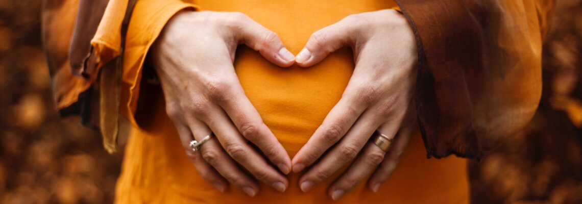 Frau hält ihre Hände geformt zu einem Herzen vor ihrem Babybauch